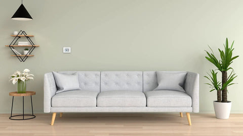 Living room with wall-mounted non programmable thermostat with backlit display and no fuss simple manual controls, precise one degree temperature control, auto heat to cool changeover, battery change reminders, white finish next to modern grey sofa and decorative plants.