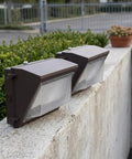 SILENRADAWN Outdoor Security Flood Lights 2 Pack Brown Aluminum Dusk to placed on a concrete wall with greenery in the background.