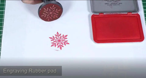 Engraving rubber stamp with red snowflake design pressed onto white paper; red ink pad case visible nearby.