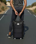 Back view of a person on a road carrying a black expandable rolling duffel bag with top handles and two wheels