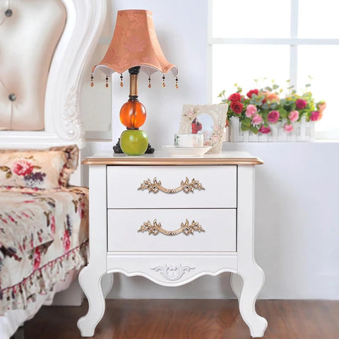 White nightstand with two drawers featuring European style vintage handles in an antique gold finish, beside a bed with floral bedding.