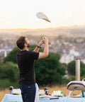 Person tossing pizza dough near Karu 12 Multi Fuel Outdoor Portable Pizza Oven delivers 950 F heat for wood or propane.