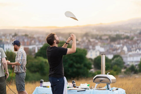 Person tossing pizza dough near Karu 12 Multi Fuel Outdoor Portable Pizza Oven delivers 950 F heat for wood or propane.