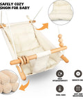 Cream-colored baby swing with cushion, wooden handles, and adjustable ropes for indoor and outdoor use with 5-point safety belt.