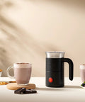 Electric Milk Frother With Handle That Delivers Barista Quality Foam From 3 To 10 Ounces In A Stainless Steel Nonstick Cup Easy Clean Compact Design With Dual Whisk For Frothing And Heating And An Ergonomic Grip displayed on a table with two mugs of frothed milk beside it.