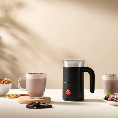 Electric Milk Frother With Handle That Delivers Barista Quality Foam From 3 To 10 Ounces In A Stainless Steel Nonstick Cup Easy Clean Compact Design With Dual Whisk For Frothing And Heating And An Ergonomic Grip displayed on a table with two mugs of frothed milk beside it.