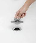 Hand placing Dual Purpose Silicone Shower Drain Stopper And Hair Catcher grey silicone over drain in bathtub.