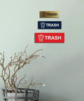 Wall display of three trash signs in different colors, including the Basic Trash Sign Brushed Silver Small For Walls Or Doors.