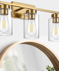 Clean Three Light Bathroom Vanity Wall Sconce In Smooth Brushed Gold Finish With Clear Retro Glass Shades above a round mirror.