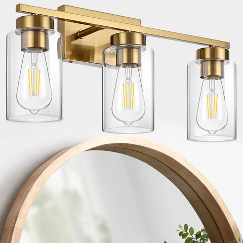Clean Three Light Bathroom Vanity Wall Sconce In Smooth Brushed Gold Finish With Clear Retro Glass Shades above a round mirror.