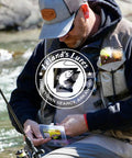 Angler holding TNT Kit 82-Piece Trout Lure Set With 70 Soft Plastic Bodies and 12 Jig Heads by a river. Fisherman gear visible.