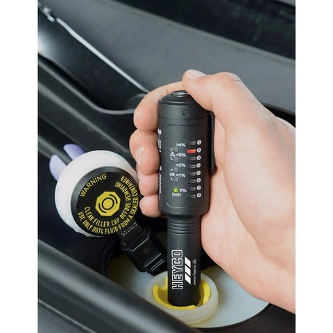 Close-up of Heyco 8-in-1 brake fluid tester, switchable, held in hand near a car brake fluid reservoir.