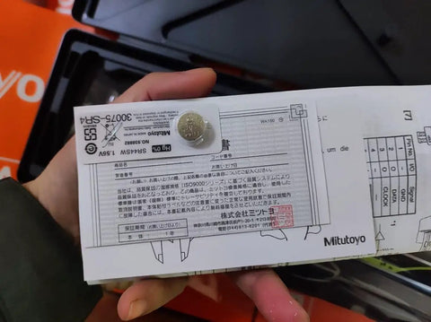 Person holding packaging for Mitutoyo product, showing specifications and a warranty card with a coin for scale