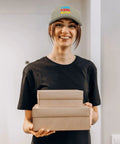 Smiling person wearing an Apricot Mesh Baseball Hat For Men And Women Cool Bugs Trucker Cap holding boxes indoors.