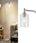 Modern metal floor lamp with clear glass shade in living room setting, adjacent to armchair and coffee table, lit bulb visible