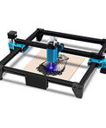 Front view of a desktop laser engraver with black frame and blue accents engraving a wood sheet.