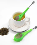 Long Handled Tea Infuser With Fine Mesh Strainer And Built In Plunger Reusable Loose Leaf Coffee Filter in green near a cup of tea.