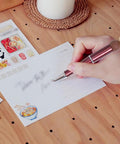 Person writing with beadable pen, next to stickers on wooden table.