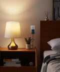 Adjustable AA Battery Power Supply Adapter plugged in powering a bedside lamp, visible on a nightstand next to a bed in a cozy room.