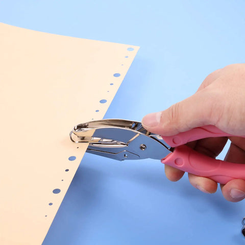 Hand using Three Piece Metal Handheld Hole Punch Set with pink plastic handle for DIY crafts on paper, showing hole creation for school supplies.