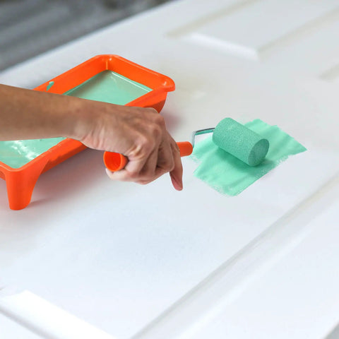 Two Inch Density Foam Paint Roller With Frame For Corner And Trim Jobs in use, applying paint on a white door with paint tray nearby.