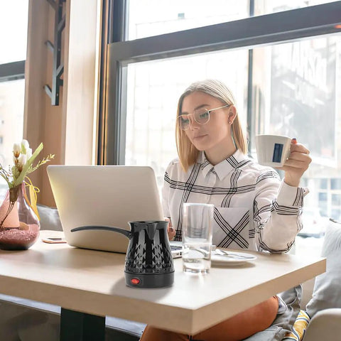 Electric Turkish Coffee Maker 500ml Pot With Removable Handle on table next to woman using laptop in a modern cafe setting