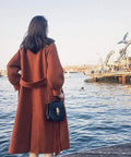 Woman in a rust-colored coat with a small bag, facing a waterfront, enjoying the view near a marina.