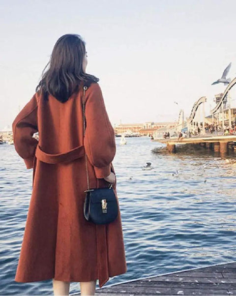 Woman in a rust-colored coat with a small bag, facing a waterfront, enjoying the view near a marina.