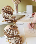 Three Dimensional Wooden Globe Puzzle Kit For Adults, Mechanical World Model, Brain Teaser And Educational Globe Build Set, DIY Wooden Globe Craft Hobby For Adults To Build And Show Off. Wooden globe puzzle on desk beside sketching hand