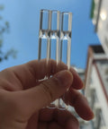 Close-up view of hand holding three 4-inch borosilicate glass tubes against a blue sky, part of Six Pack Borosilicate Glass Tubes For Lab And DIY Blowing, 4 Inch Length, 12 Mm Outer Diameter, 2 Mm Thick Wall, High Heat And Chemical Resistant, Premium Glass Tubes For Science Projects, Glassblowing And Crafting.