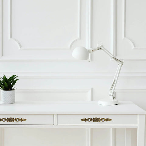 White desk with Vintage Bronze Patina Finish Cabinet Hardware Set floral pulls, lamp, and plant, in bright room with paneled wall.
