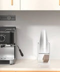 Two Tier Upright Cup And Condiment Organizer With Two Compartment Clear Acrylic Cup Dispenser, Straw Holder And Lid Storage for breakroom coffee station next to espresso machine.