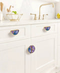 Two Pack Fluid Art Cabinet Pulls in Purple Teal Gold on white kitchen cabinets with gold semicircle design and marble-like pattern.