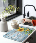 Person placing cup on MustHave Kitchen Dish Drying Mat 18x24 with watercolor sunflower on gradient blue wood grain countertop
