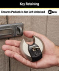Hand holding Beefed Up Stainless Steel Discus Lock with Tough Cut Boron Carbide Shackle near a latch, ideal for security.
