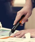 REXBETI retractable utility knife with orange thumb slider cutting cardboard.
