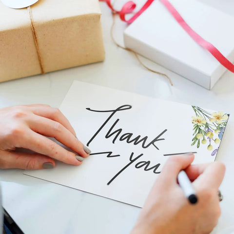 Person writing on 100 Piece Floral Blank Note Cards Set, 4 by 6 Inches, with floral corner, suitable for greeting cards, white background.