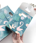 Hands holding box with flamingo design, floral patterns, and plants on a teal background. Box has punch-out handle.