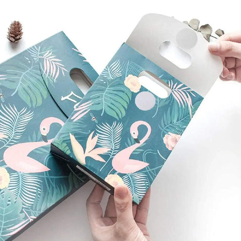 Hands holding box with flamingo design, floral patterns, and plants on a teal background. Box has punch-out handle.