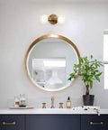Rock This Mini Two Light Gold Bathroom Vanity Wall Sconce With Clear Glass Shades installed above round mirror in modern bathroom.