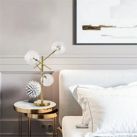 Modern Gold Table Lamp With 3 Light Glass Globe Bulbs, Antique Brass Finish on a nightstand by a bed in a bedroom setting.