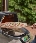 Karu 2 Pro Multi Fuel Pizza Oven For Backyard Grilling Wood Or Gas Fired 16 Inch Stone Pizza Maker with pizza being placed inside.