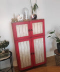 Vintage red cabinet with white panels, featuring small black ring knobs, surrounded by indoor plants and decor items.