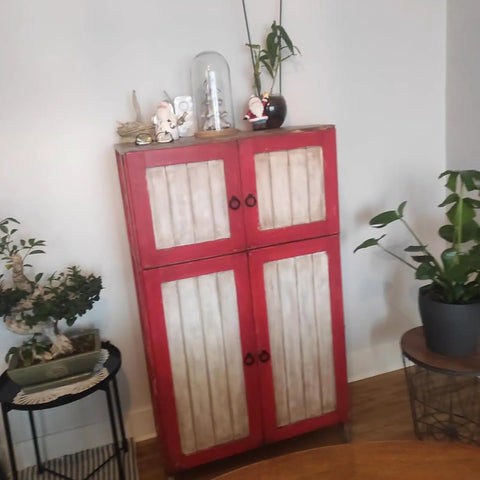 Vintage red cabinet with white panels, featuring small black ring knobs, surrounded by indoor plants and decor items.