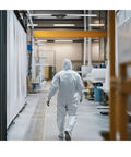 Person wearing Heavy Duty Hazmat Coveralls 25 Pack With Hood And Elastic Wrists, Full Body Disposable Suit in a warehouse setting.