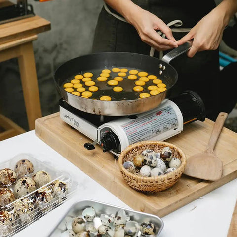 Person cooking quail eggs in a pan with 50 Pack Quail Egg Cartons nearby, showcasing eggs stored for easy transport in clear PVC trays.