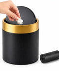 Hand using Mini Desktop Trash Can With Swing Lid Sleek Black And Brushed Gold Rim 1.5 Liter Capacity Compact 5.2 Inch Tall And 4.7 Inch Diameter Great For Desk Vanity Bathroom Coffee Bar And Office