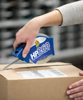 Person using high performance clear packing tape dispenser on box, demonstrating all weather grip and super strong adhesive.