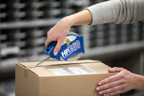 Person using high performance clear packing tape dispenser on box, demonstrating all weather grip and super strong adhesive.