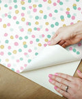 Hands peeling Pink And Blue Glitter Confetti Peel And Stick Wallpaper Sample, showing repositionable and removable feature on wood surface.